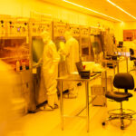 Workers in lab coats and safety gear working in the yellow-tinted clean room.