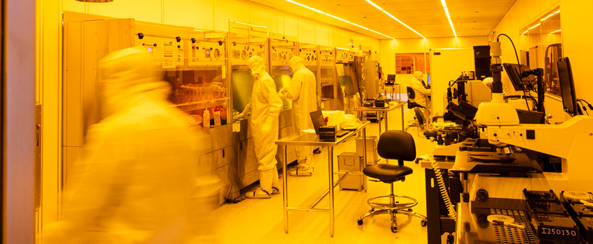 Workers in lab coats and safety gear working in the yellow-tinted clean room.